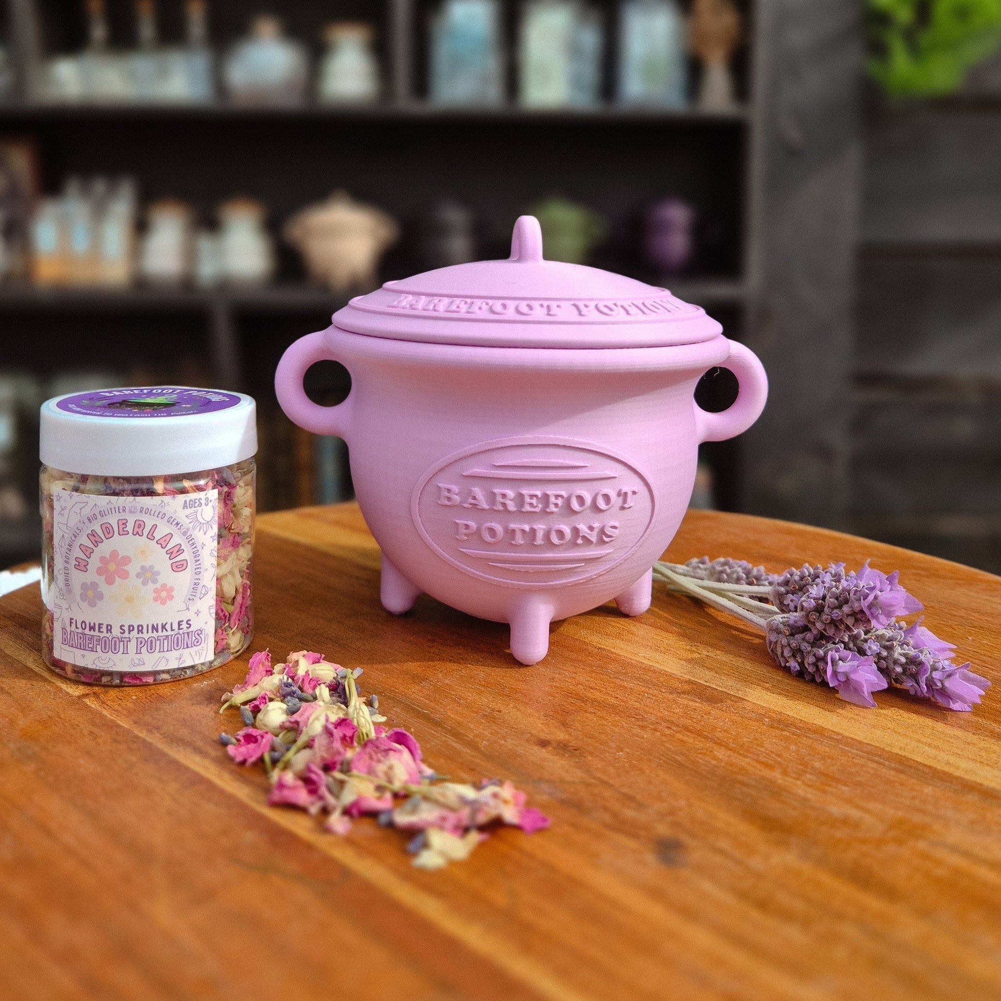 Purple cauldron with 'Barefoot Potion' branding on a wooden surface, surrounded by dried flowers and a jar.