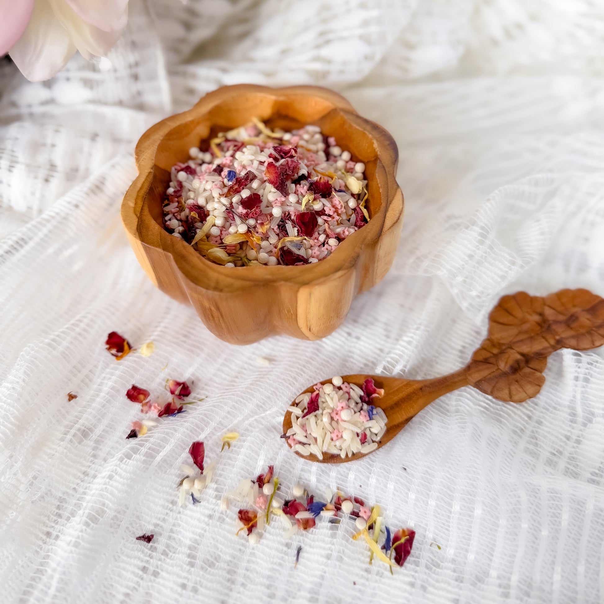 Wooden bowl and spoon with fairy sensory base including dried flowers, pink grains and rice on a textured white fabric background. Used for nature and water play with children, sensory exploration.