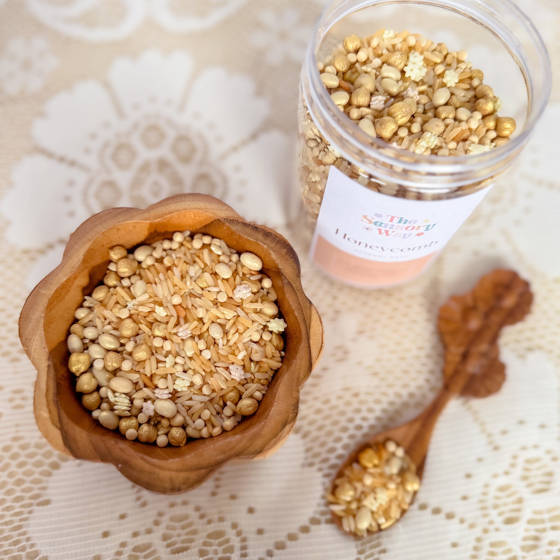 Wooden bowl and jar filled with golden seeds and rice sensory base on a lace tablecloth. Used for nature play with children, sensory exploration.