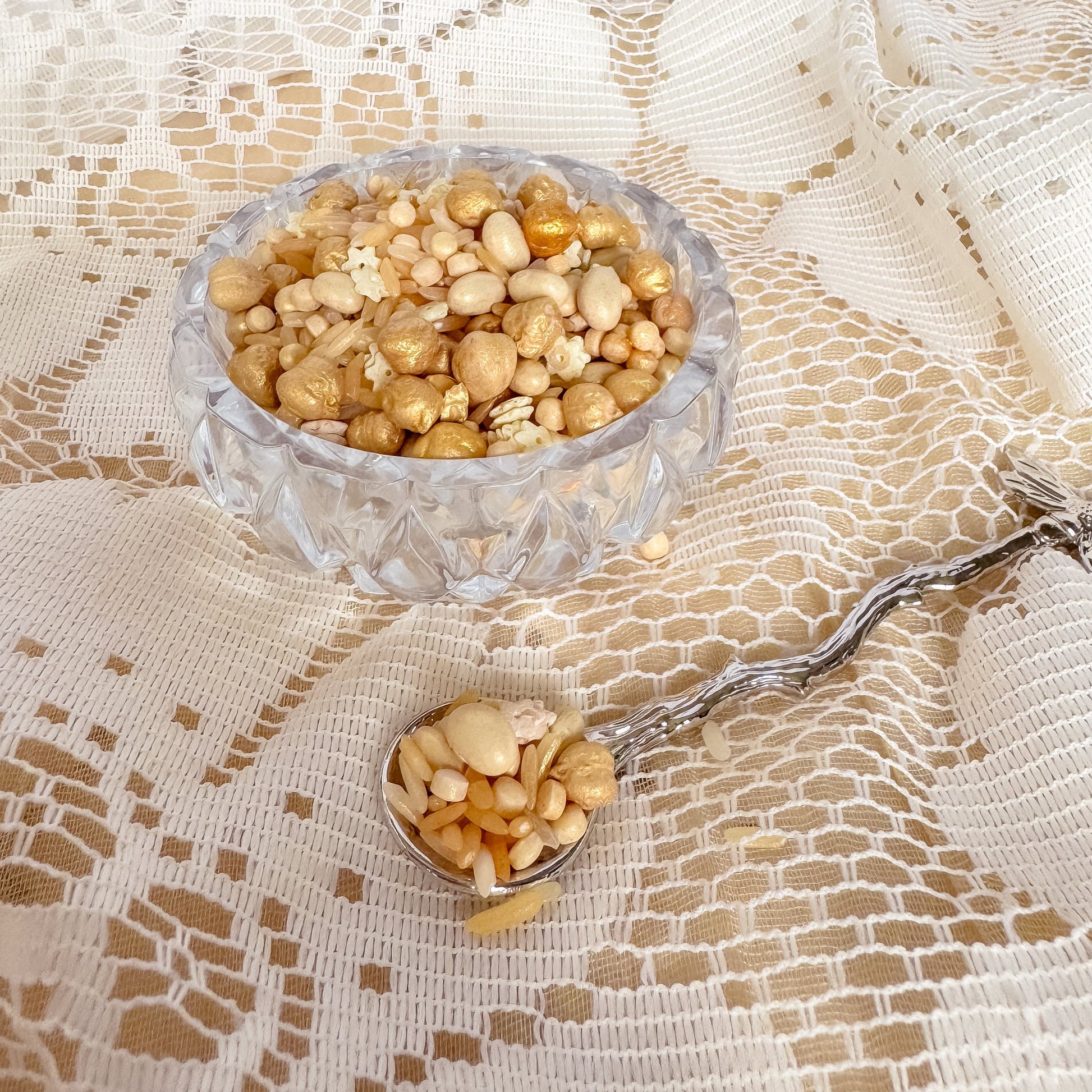 Clear glass bowl and spoon filled with golden sensory base including rice, grains and chickpeas on a textured white fabric background