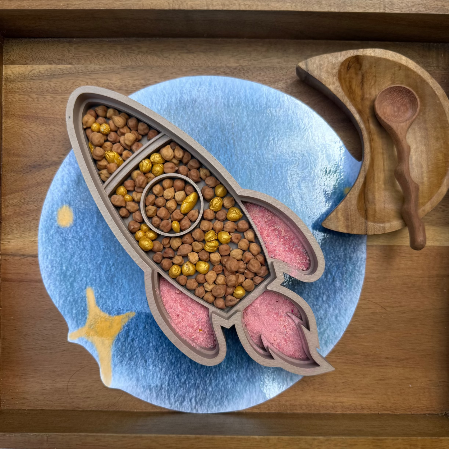 Rocket shaped sensory tray, with chickpea sensory mix and fizz, wooden spoon on the side, on a wooden bin. Moon bowl.