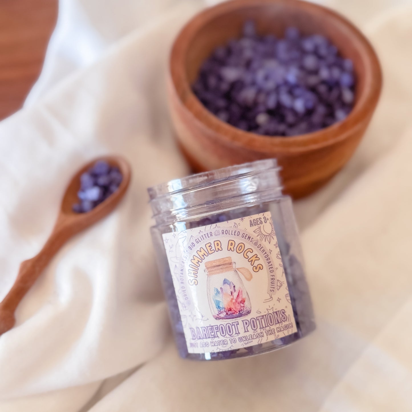 Jar of purple shimmer rocks with a wooden bowl and spoon on a white fabric background, for potion play, sensory
