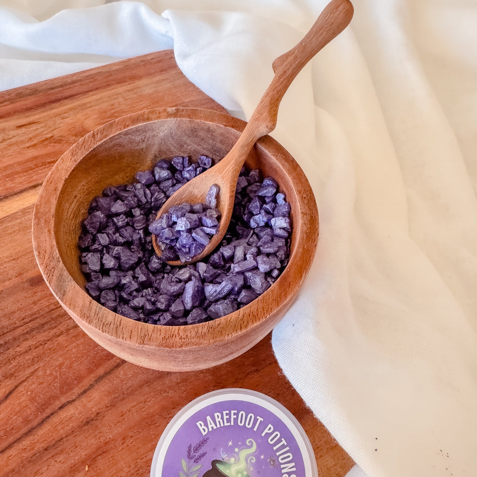 Wooden bowl with purple shimmer rocks and a wooden spoon on a wooden surface, with 'Barefoot Potions' product below.