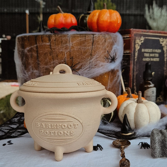 Decorative beige cauldron labeled 'Barfoot Potions' on a Halloween-themed setting with pumpkins and cobwebs.