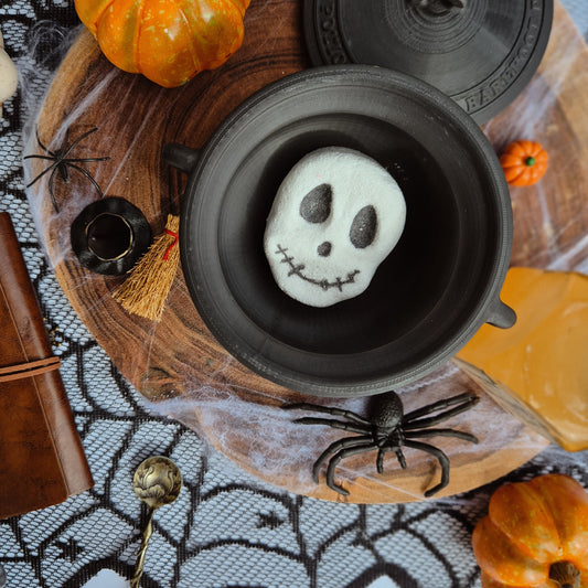 Halloween-themed setting with a skull-shaped fizzer in a black bowl, pumpkins, and spider decorations on a lace tablecloth.