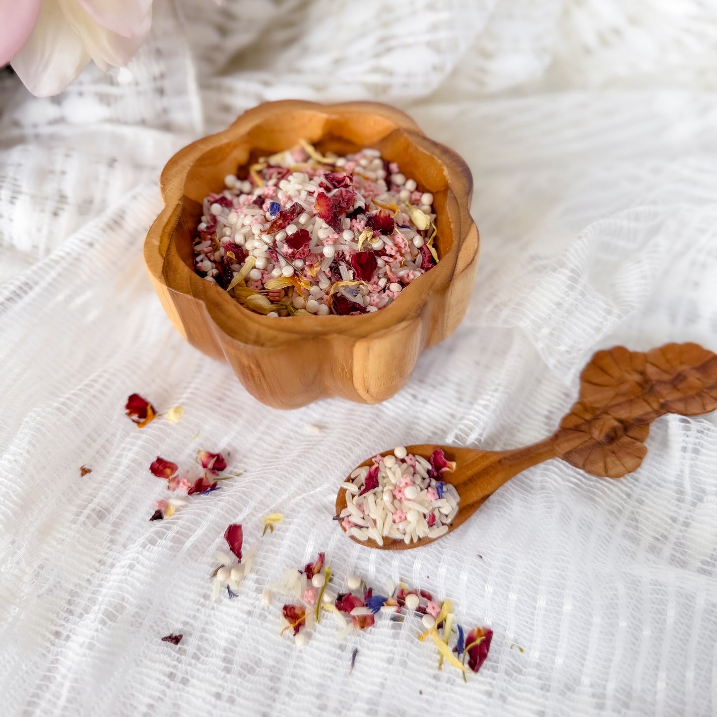 Wooden bowl and spoon with fairy sensory base including dried flowers, pink grains and rice on a textured white fabric background. Used for nature and water play with children, sensory exploration. 