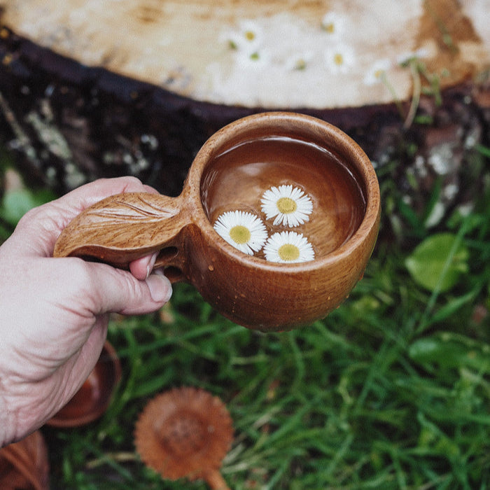 Wooden mug with leaf shaped handle, flowers in water, held by a hand against a natural background. Used for nature and water play with children, sensory exploration. 