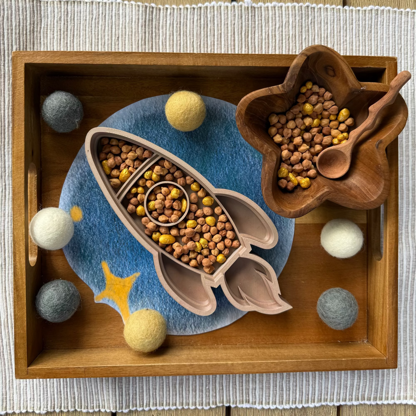Wooden tray with decorative elements resembling a rocket and celestial bodies on a wooden surface. Rocket shaped sensory tray, with chickpea sensory mix and fizz, wooden spoon on the side, on a wooden bin. Moon bowl. 