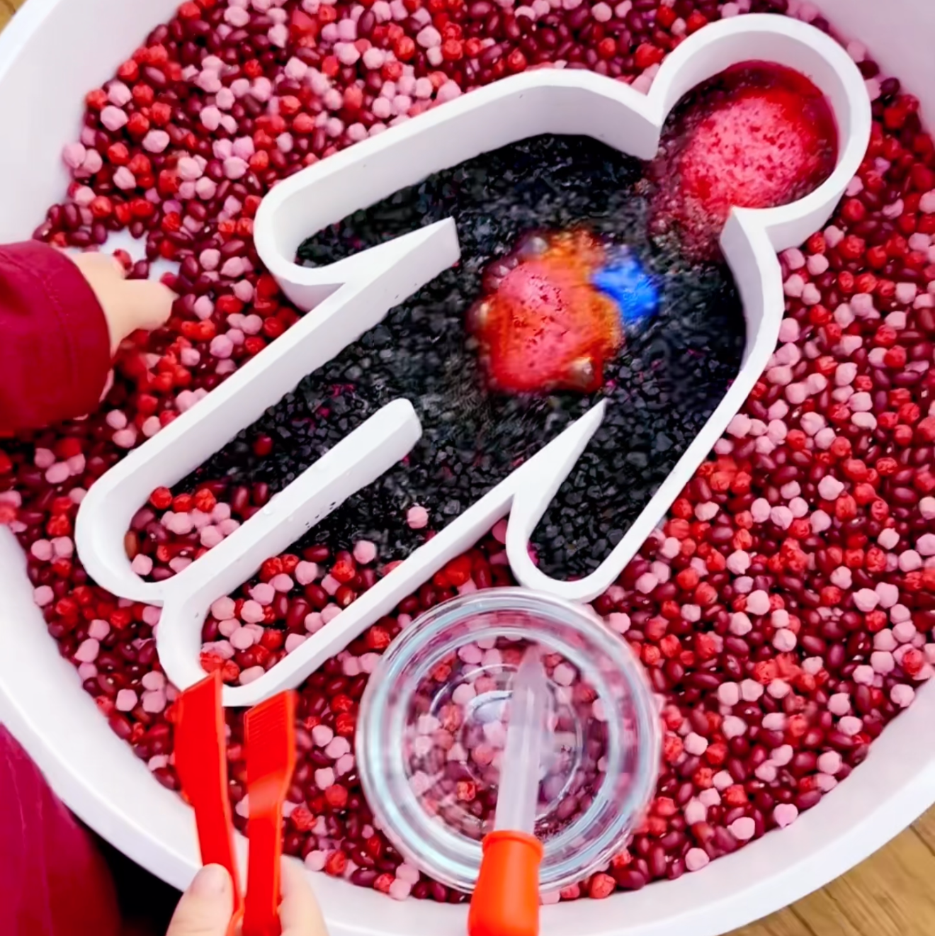 Child playing with a sensory bin filled with red and black beads, using a scoop to interact with the contents.