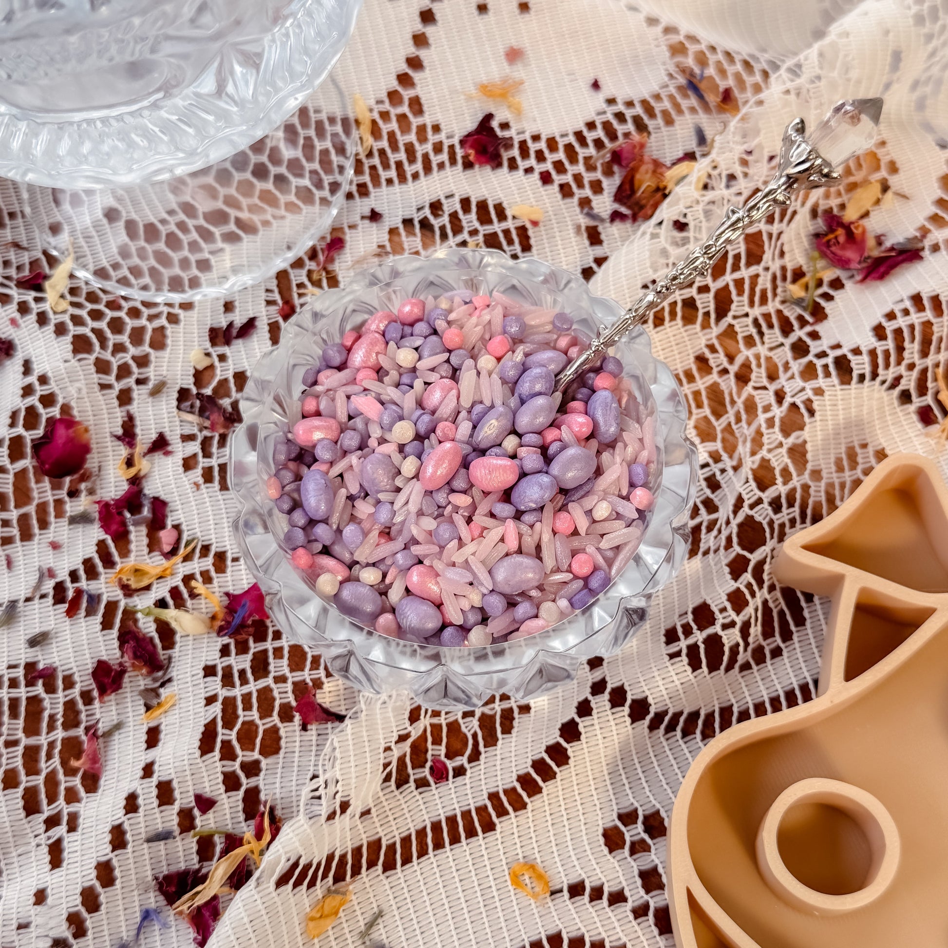 Decorative glass bowl with pink and purple sensory base made up of rice and grains, on a lace tablecloth. Used for nature and water play with children, sensory exploration. 
